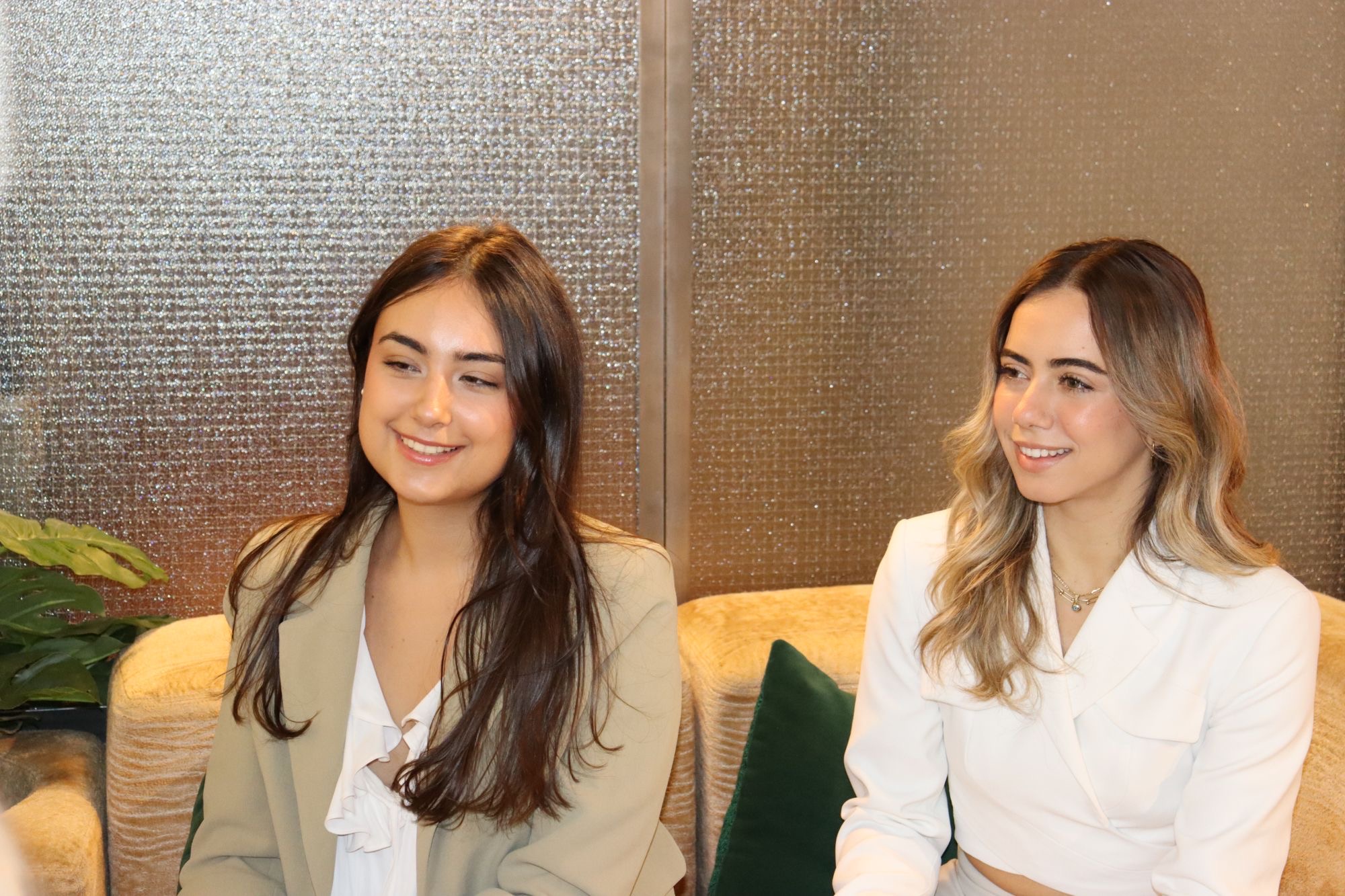 Two women in blazers sit close together on a golden sofa, smiling as they look away from the camera