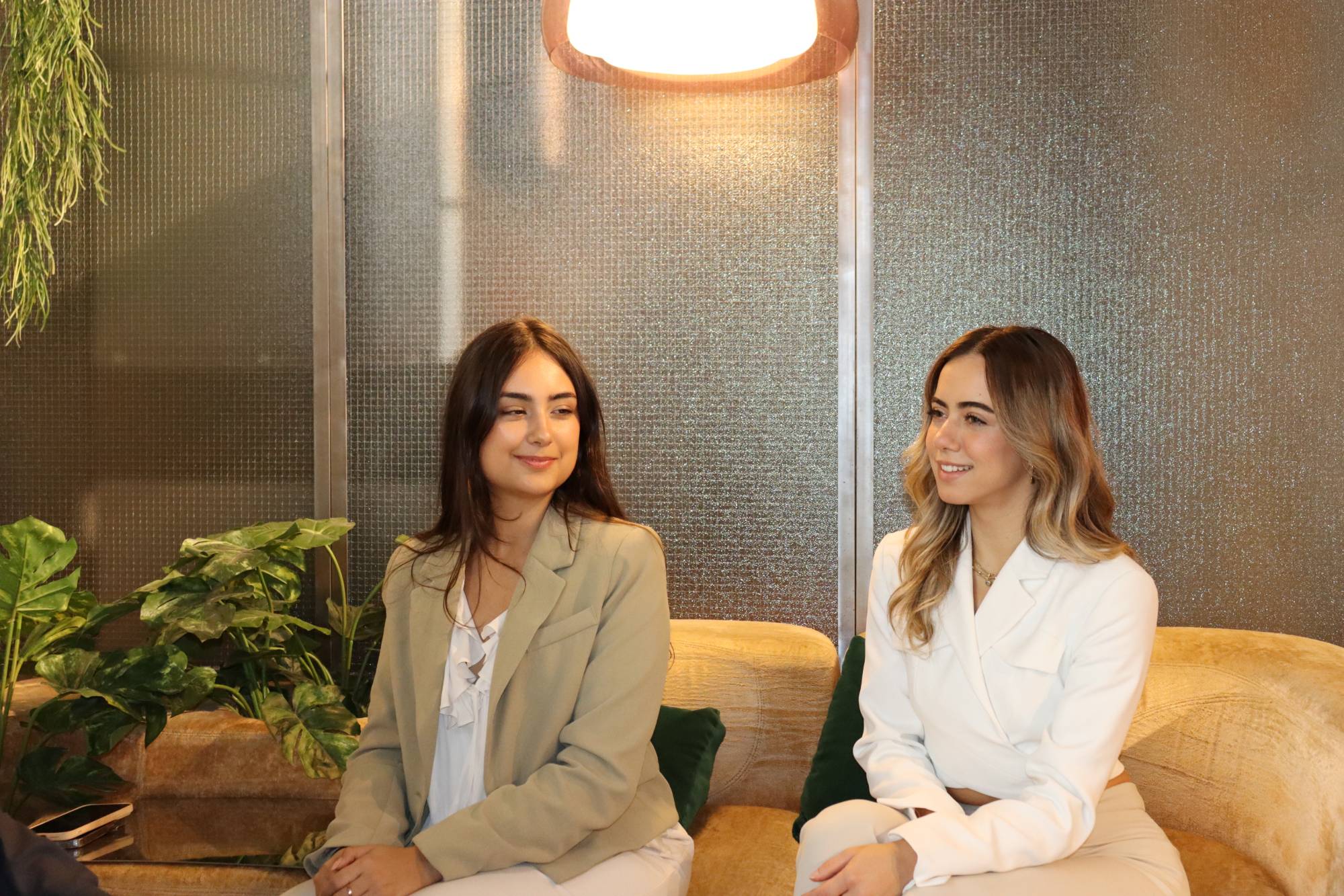 Two women in blazers sit side by side on a golden sofa under a warm lamp, smiling as they listen to someone out of frame